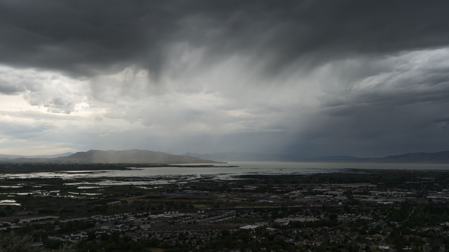 A rainy landscape of lake, mountains, and town with a progression from right to left where it's darker than lighter also the sheet of rain muddies the color of the landscape behind just slightly